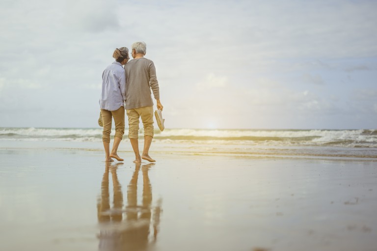 Senior Couple Walking On The Beach Holding Hands At Sunrise
